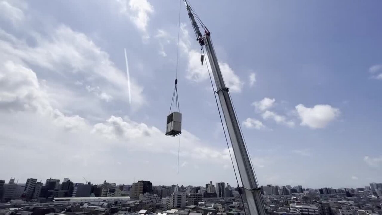 クレーンで吊り下げられた空調室外機が、青空を背景にゆっくり降りてくる様子